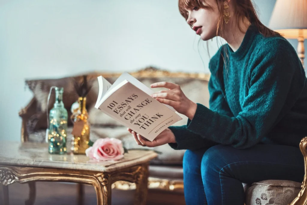 Woman sitting on a vintage sofa reading a book titled '101 Essays That Will Change the Way You Think' with decorative bottles and a pink rose on a gold table nearby